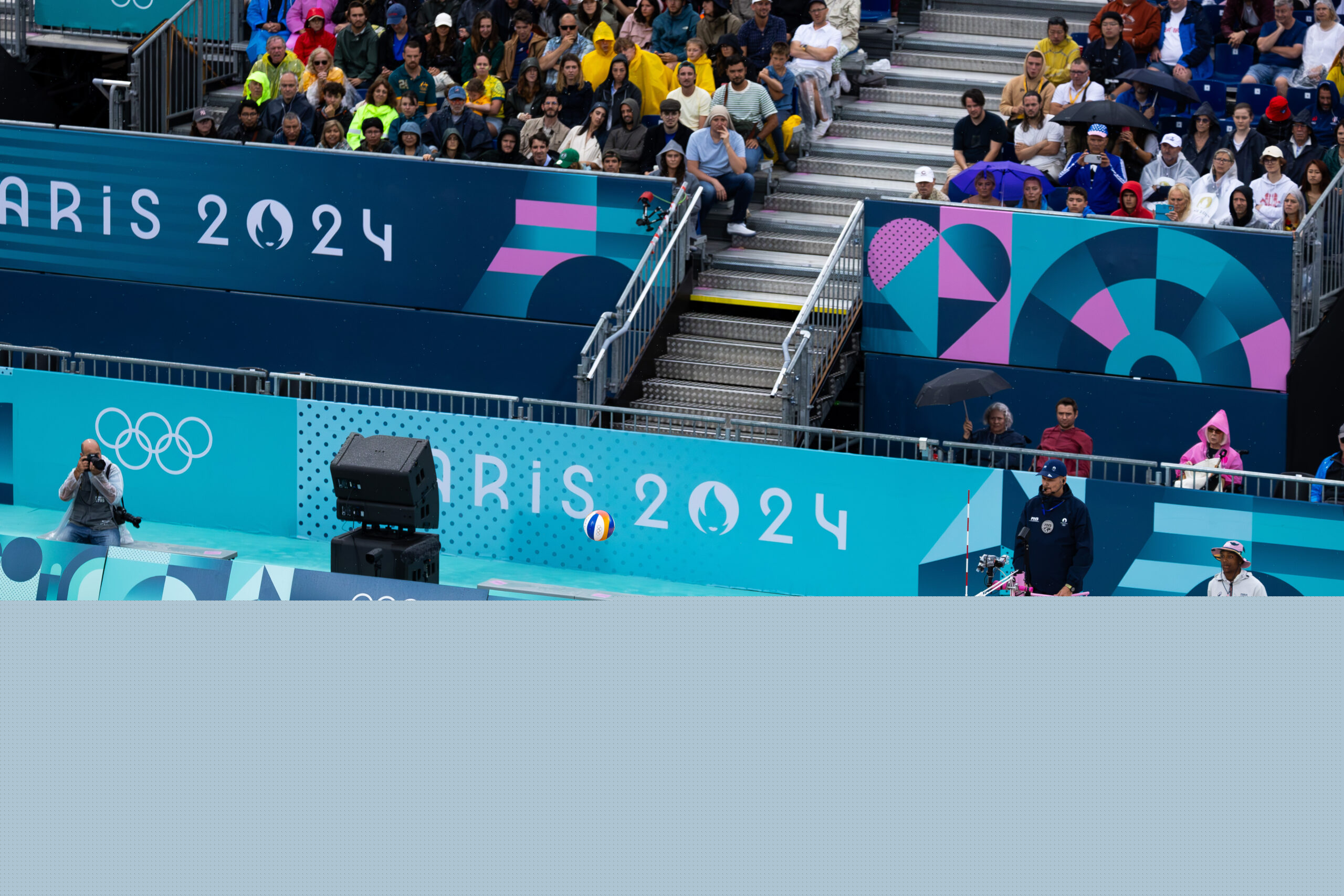 Mens beach volleyball at the Olympics. A player in red leaps into the air preparing to hit the ball. Another player in yellow on the other side of the year jumps with his hands in the air to block the oncoming shot. Photographers and a crowd on a stand are in the background.